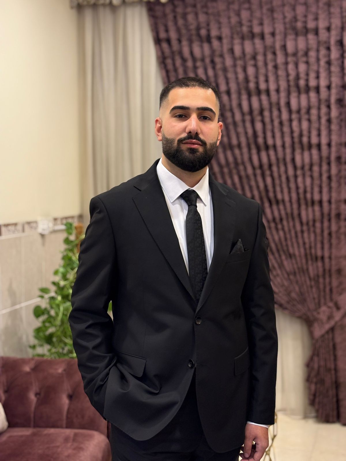 Portrait photo of Yousif Munir Butrus wearing a black suit, white shirt, and black tie standing indoors